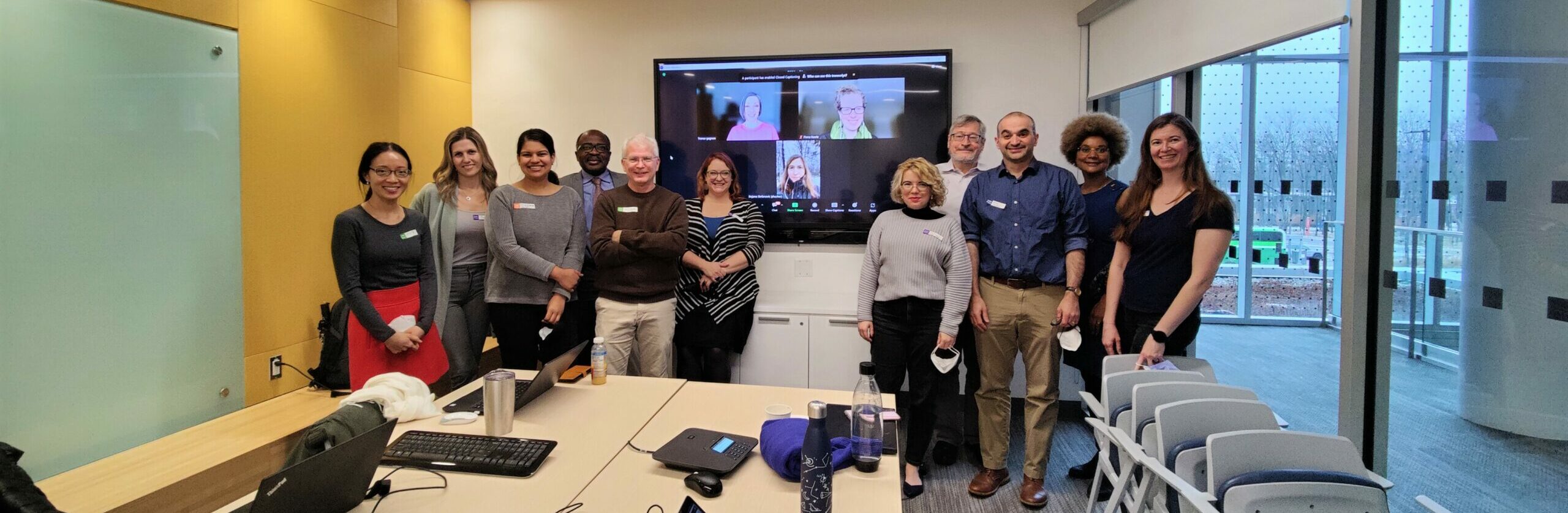 A group of people stand smiling at the end of a conference table in a room with one wall-sized window. A screen in the middle of the group shows three people online.