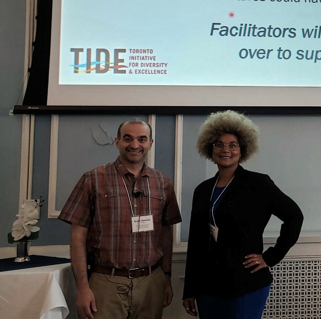 Two people pose in front of a projection screen at an in-person TIDE session